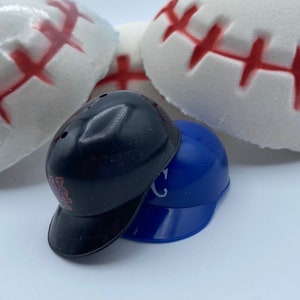 May include: A close-up shot features two miniature baseball helmets, one black and one blue, stacked together. The black helmet has the letters "Mets" on the side. Behind them are several white baseball-shaped items with red stitching.