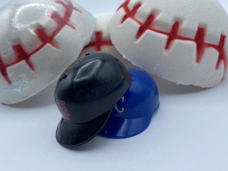 May include: A close-up shot features two miniature baseball helmets, one black and one blue, stacked together. The black helmet has the letters "Mets" on the side. Behind them are several white baseball-shaped items with red stitching.