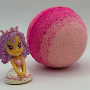 May include: A small princess figurine with a purple crown, hair, and a white dress with pink trim. A large, pink bath bomb with glitter sits next to the figurine. The bath bomb is a gradient of light and dark pink.