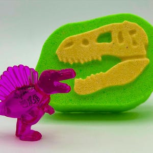 May include: A translucent magenta dinosaur toy with a spiky back fin stands next to a green sponge with a yellow dinosaur skull imprint. The toy has the letters "KCSB" on its side. The skull imprint is a fossil-like design.