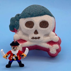 May include: A skull and crossbones bath bomb with a teal bandana, white skull, and red base, positioned behind a small plastic pirate figurine wearing a red coat, black hat, and eye patch, holding two white swords in its hands.