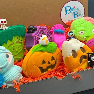 May include: A cardboard box filled with orange paper shreds and various Halloween-themed bath bombs, including shapes of a green monster, a purple spiderweb, a pumpkin, a candy corn, and a mummy, with small plastic figures like a skeleton and reaper.