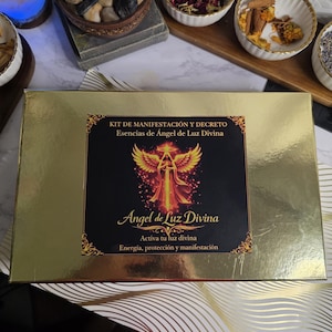 Puede incluir: Una caja dorada con el texto "KIT DE MANIFESTACIÓN Y DECRETO Esencias de Ángel de Luz Divina" y una ilustración de un ángel. La caja está sobre una superficie blanca con pequeños cuencos de hierbas y especias secas.