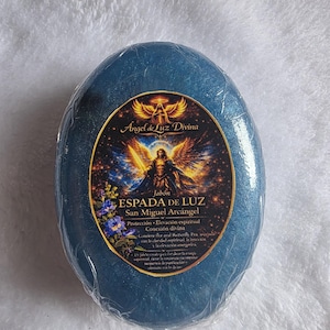 Puede incluir: Pastilla de jabón azul ovalada con una etiqueta que presenta una ilustración de un ángel y el texto "ESPADA DE LUZ San Miguel Arcángel". El jabón está envuelto en plástico transparente. La etiqueta también incluye el texto "Angel de Luz Divina".