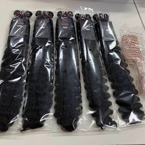 Könnte beinhalten: Fünf Bündel schwarzer, lockiger Haarverlängerungen in durchsichtiger Plastikverpackung, zusammen mit einer rosa Haarbürste. Die Haarverlängerungen sind fest gebündelt und mit einem roten Faden zusammengebunden. Die Haarbürste befindet sich ebenfalls in einer transparenten Verpackung.