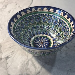 May include: A ceramic bowl with a detailed floral pattern in shades of blue, green, and white. The bowl has a dark blue rim and a decorative design on the inner surface. The bowl is likely used for serving food or as a decorative piece.