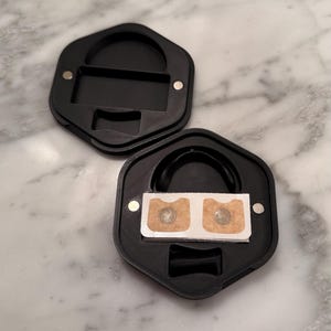 May include: Two black, hexagonal cases, one open and one closed, with magnetic closures. The open case reveals a black curved object and a white rectangle with two circular, metallic objects. The cases are on a marble surface.