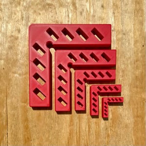 Puede incluir: Un conjunto de cinco escuadras de sujeción en forma de L de color rojo, encajadas entre sí. Cada escuadra presenta una serie de recortes rectangulares diagonales a lo largo de ambos brazos, creando un patrón geométrico uniforme en todos los tamaños.