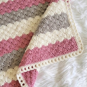 May include: A crochet blanket featuring a repeating chevron pattern in alternating stripes of dusty rose pink, light gray, and cream white. The edges are finished with a decorative cream-colored border consisting of small, open square cutouts.
