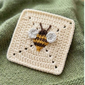 May include: A square, cream-colored crochet piece featuring a detailed bee applique. The bee has brown, yellow, and gray accents, with small black dots around it. The piece is set against a green knitted background.