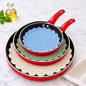 May include: Two red frying pans stacked with three round crochet doilies. A light blue doily sits inside the smaller pan, a sage green doily sits inside the larger pan, and a cream doily sits under the larger pan.