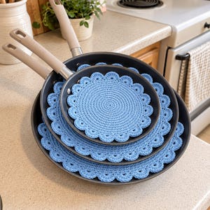 May include: Two dark gray non-stick frying pans with light beige handles are stacked together. Each pan contains a round, light blue crocheted mat with a scalloped edge, designed to protect the interior surfaces from scratches.
