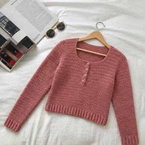 Può includere: Un maglione a maniche lunghe color rosa polvere realizzato con un motivo a uncinetto. Il capo presenta uno scollo quadrato, un orlo a coste, polsini a coste e un'abbottonatura frontale fissata da quattro bottoni rotondi abbinati su gruccia.