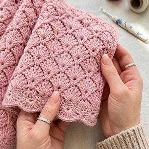 May include: A close-up of a handmade, light pink crocheted item. The intricate pattern features a repeating fan-like design. The edges have a scalloped border. The item is held by two hands, showcasing the texture and detail of the craft.