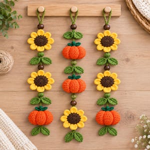 May include: Three vertical hanging wall decorations made of crocheted sunflowers and orange pumpkins. Each strand features alternating yellow and brown flowers, orange pumpkins, green leaves, and dark brown wooden beads on a green yarn loop.