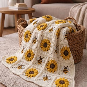 Puede incluir: Una manta de ganchillo con un patrón cuadrado repetitivo de girasoles amarillos con centros marrones y pequeñas abejas con alas blancas, sobre un fondo blanco roto. La manta está colocada sobre una cesta de mimbre marrón.
