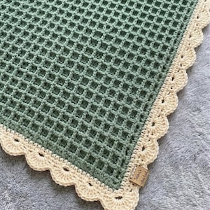 Puede incluir: Una manta de ganchillo verde salvia hecha a mano con un patrón de punto de gofre. La manta tiene un borde festoneado de color crema. Una pequeña etiqueta rectangular con texto está adherida a la manta.