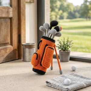 Può includere: Una sacca da golf all'uncinetto in arancione brillante e nero, dotata di supporto e tasche esterne. All'interno della sacca si trovano diverse mazze da golf all'uncinetto nei toni del grigio, nero e arancione. Due palline da golf bianche a terra.