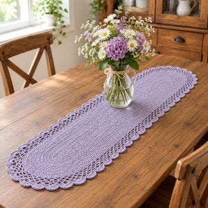 May include: An oval-shaped, light purple crochet table runner featuring a decorative scalloped edge. The runner is placed on a wooden table and supports a clear glass vase filled with a bouquet of white daisies and purple hydrangea flowers.