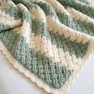 May include: A handmade crochet blanket in a basketweave pattern. The blanket features alternating stripes of sage green and cream-colored yarn. The edges are finished with a scalloped cream border, creating a cozy and textured appearance.