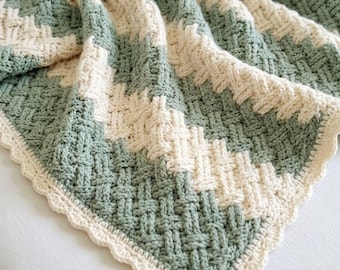 May include: A crochet blanket featuring a basketweave texture with diagonal stripes in sage green and cream. The edges are finished with a scalloped border in cream, creating a soft, woven appearance throughout the entire textile piece.