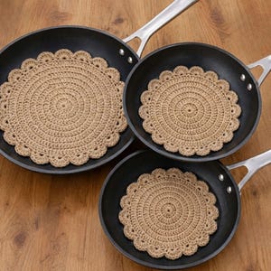 May include: Three round, beige, crocheted fabric pads with scalloped edges are placed inside three black non-stick frying pans of varying sizes. Each pad features a circular, textured pattern made of yarn, designed to fit within the pans.