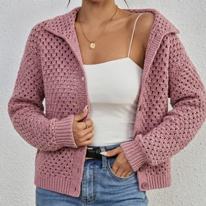 May include: A dusty rose crochet cardigan featuring a collared neckline, long sleeves with ribbed cuffs, and a button-down front. The garment is worn over a white camisole and blue denim jeans, accessorized with a gold pendant necklace.