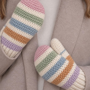 May include: A pair of crochet mittens featuring horizontal stripes in pastel pink, sage green, light blue, tan, and lavender, separated by thin cream-colored bands. The mittens have a ribbed cream cuff and a thumb section on the right side.