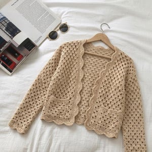 May include: A beige crochet cardigan featuring a granny square pattern, long sleeves, two front pockets, and a scalloped edge along the open front and neckline. The garment is displayed on a wooden hanger, showcasing the intricate openwork knit texture.
