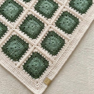 May include: A crocheted blanket with a repeating pattern of green squares set within off-white borders. The blanket has a textured finish and a tag is attached to the corner. The colour scheme is neutral with a hint of green.