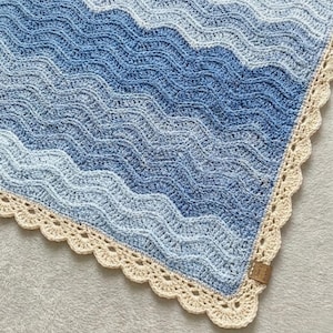 May include: A handmade crochet blanket with a wave pattern in shades of blue and white. The blanket has a scalloped, cream-colored border. A small, brown tag is attached to the corner. The blanket is a cozy, decorative item.