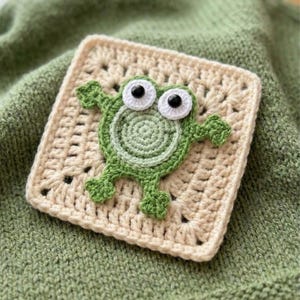 May include: A crocheted square with a green frog applique. The frog has large white eyes with black pupils and is centered on a cream-colored square. The background is a soft green fabric.