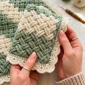 May include: A handmade crochet blanket with a basketweave design. The blanket uses sage green and cream yarn in a woven pattern. The edges are finished with a scalloped border. A crochet hook and yarn are visible in the background.