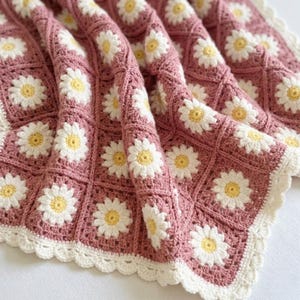 May include: A handmade crochet blanket in a dusty rose colour, featuring a repeating pattern of white daisies with yellow centres. The blanket has a scalloped white border, creating a cosy and decorative textile.