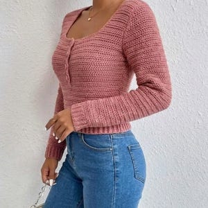 May include: A dusty rose long-sleeve crochet top featuring a square neckline, a button-down placket with three matching buttons, and ribbed cuffs. The garment is paired with blue denim jeans, creating a textured and fitted appearance.