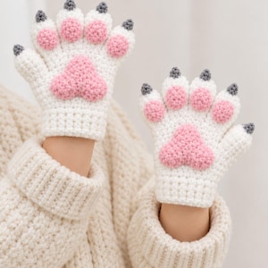 May include: A pair of white crochet fingerless gloves designed to resemble animal paws. Each glove features four small pink circular pads on the fingers, a larger central pink pad on the palm, and dark grey textured tips on each finger.