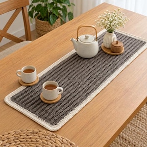 Op de afbeelding: Een rechthoekige donkergrijze gehaakte tafelloper met een dikke crèmekleurige rand. De loper ligt op een houten tafel en bevat een witte keramische theepot, twee witte keramische mokken op houten onderzetters en een kleine witte vaas met bloemen.
