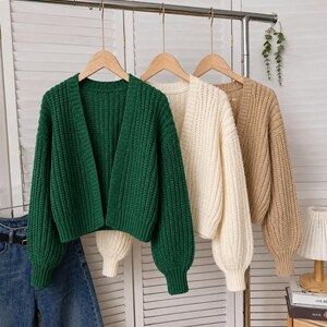 May include: Three open-front cardigans hanging on wooden hangers. The cardigans feature a chunky ribbed knit texture, long sleeves with ribbed cuffs, and a V-neckline. The colors are forest green, cream, and tan, each with a cropped length.