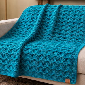 May include: A teal-colored, textured crochet blanket draped over a beige sofa. The blanket features a repeating pattern of raised, diamond-shaped designs. A small, rectangular leather tag is sewn on the bottom right corner of the blanket.