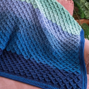May include: A handmade crochet blanket in shades of blue and green. The blanket features a textured, scale-like pattern, with a dark blue border. The colors transition from light green to deep blue, creating a gradient effect. The blanket is draped over a pink surface.