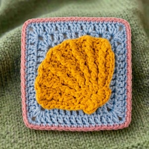 May include: A square crochet piece featuring a yellow seashell design on a light blue background, framed by a pink border. The piece is set against a green knitted surface, showcasing the handmade texture.
