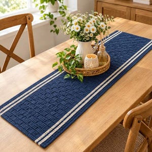 Op de afbeelding: Een rechthoekige marineblauwe tafelloper met een getextureerd mandvlechtpatroon en twee parallelle witte strepen langs elke lange zijde. De loper ligt plat op een houten tafel en vormt de basis voor een witte kan en een ronde schaal.