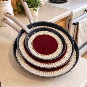 May include: Two dark gray non-stick frying pans stacked together, each featuring a round, handmade crochet pan protector. The protectors have a deep red center, a thin gold border, and a cream-colored fringe edge.