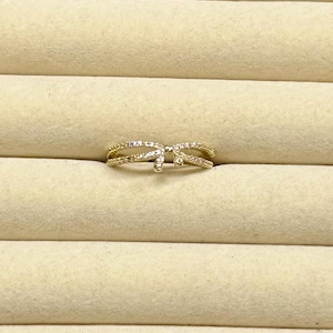 May include: A gold-colored ring with a bow design. The ring features a double band, with the top band adorned with small, clear gemstones. The bow's center is also embellished with gemstones, creating a sparkling effect.