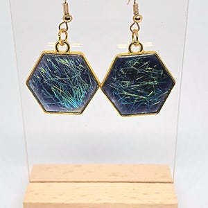 May include: A pair of gold-coloured earrings featuring hexagonal pendants with iridescent blue and green hues. The earrings have gold-coloured hooks and are displayed on a clear stand with a wooden base. The pendants have gold-coloured borders.