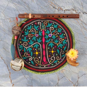 May include: A collection of spiritual items on a marble surface. Includes a wooden flute, a round black embroidered mat with a tree design, a rattle, feathers, a small bowl with stones, and a dried flower.