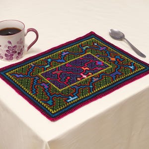 Op de afbeelding: Een kleurrijke, handgestikte placemat met een geometrisch ontwerp in groen, paars, blauw en oranje. Een koffiemok en een lepel liggen op een wit tafelkleed naast de placemat. De placemat heeft een magenta rand.