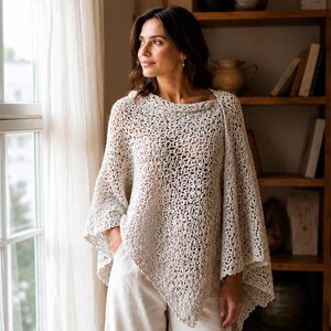 Può includere: Un poncho all'uncinetto color crema a trama aperta, con collo a imbuto e orlo asimmetrico. Il poncho è leggero e sembra fatto a mano, adatto per la stratificazione. La modella indossa il poncho con pantaloni bianchi.
