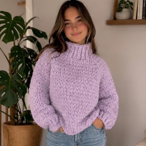 Puede incluir: Un suéter de punto grueso de color lila claro con cuello alto y acanalado. El suéter tiene un corte holgado y mangas largas. La modelo lleva vaqueros azules. El fondo incluye una gran planta en maceta y una estantería con libros.