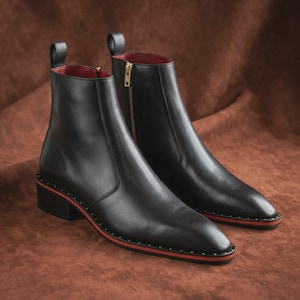 May include: A pair of black leather ankle boots with a red trim and gold zipper. The boots have a low heel and a pull tab at the back. The boots are approximately 6 inches tall.